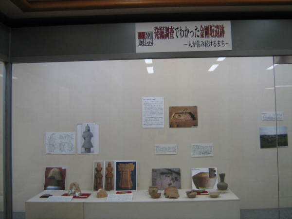 後方の壁に発掘場所などの写真が貼られ展示台に壺や杯、埴輪などが並んでいる写真