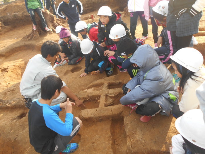 遺跡の中に子ども達が入り、十字型に土が掘り起こされた場所に子ども達が集まり、職員の男性の説明を興味深く聞いている様子の写真
