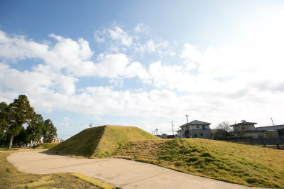 周りに小さな森や住宅地が建つ公園敷地内に台形に盛り上がった古墳の写真