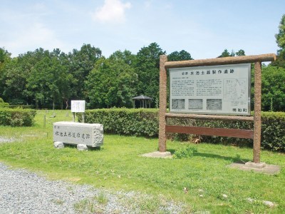 木々に囲まれた公園の生垣の手前左に横長の石造りの看板、右側に大きな遺跡の説明が書かれた看板が設置された写真