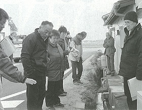 左に道路右に民家が建っている間の溝を挟んで議員の方々が集まり担当者から話を聞いている白黒写真