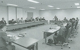 長机をロの字に設置した室内で奥の立っている男性の話を参加者が聞いている白黒写真