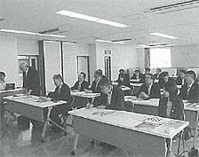 長机が2列に並んだ室内で左手前の席の男性が立ち意見を述べている白黒写真