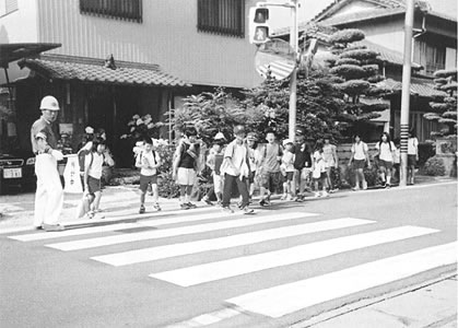 横断歩道を渡っている子供たちと旗を持っている見守りの男性の写真