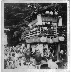 神社の境内に停まっている山車を大勢の見物客が見ている白黒写真