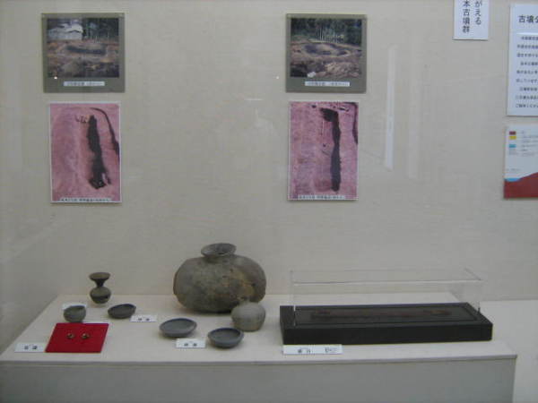 後方の壁に4枚の発掘場所の写真が貼られ、展示台に横長の瓶や杯、耳環などが展示されている写真