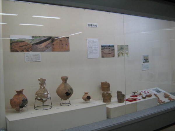 古墳時代と書かれた後方の壁に発掘場所の写真などが貼られ、手前に様々な形の壺や埴輪などが展示されている写真