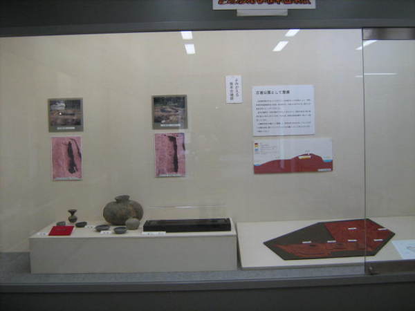 後方の壁に発掘場所の写真や地表図が貼られ、手前に横長の瓶や耳環、発掘場所の模型などが展示されている写真