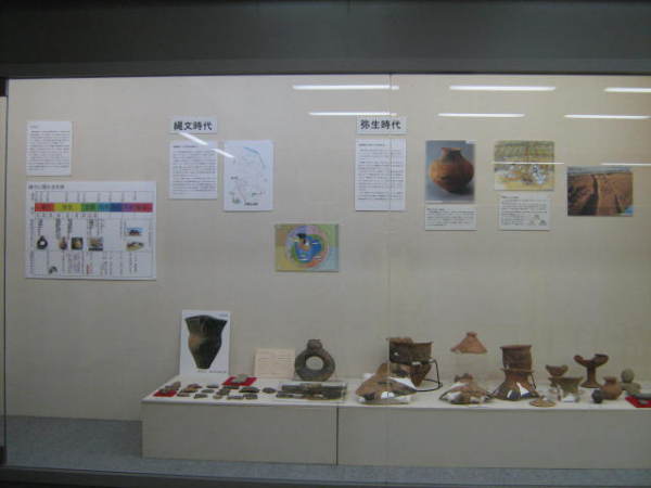 縄文時代と弥生時代と書かれた壁に写真や年表などが貼られ、手前に埴輪や土器などが展示されている写真