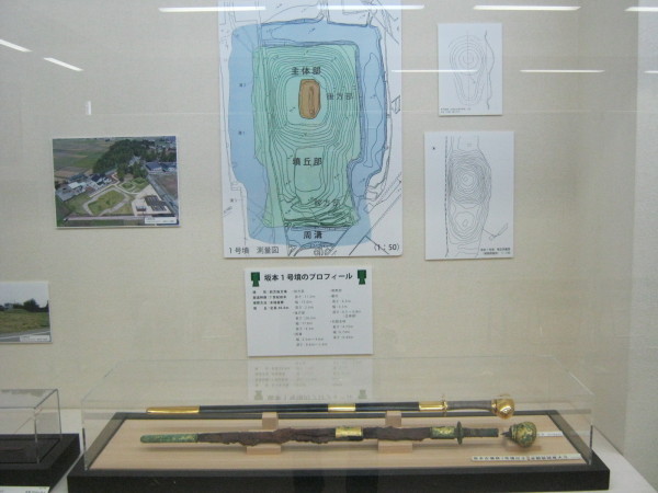 後方の壁に発掘場所の写真や図が貼られ、手前の展示台に2本の刀が展示されている写真