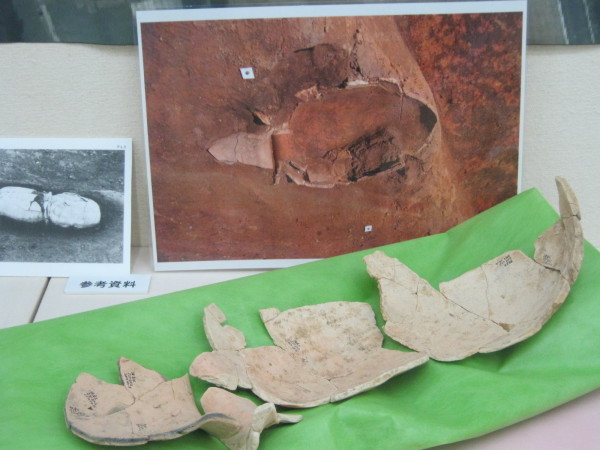 発掘場所の大きな写真と白黒写真が立てかけられ、手前の黄緑色の布の上に発掘された土器棺の欠片が並んで置かれている写真