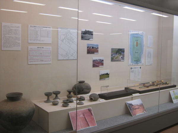 後方の壁に写真や発掘場所を上から見た図が貼られ、大きさの違う瓶や杯、刀などの展示を左斜めから撮影した写真