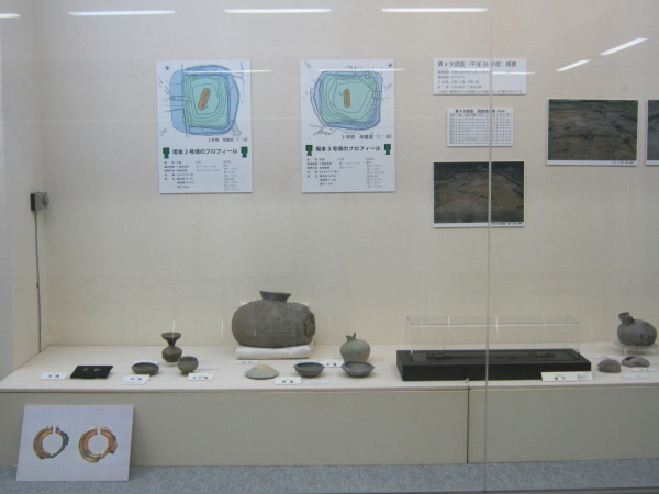 後方の壁に写真や発掘場所を上から見た図が貼られ、手前の展示台に様々な形をした瓶や刀などが展示されている写真