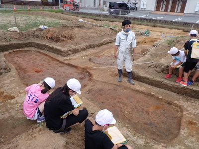 楕円形の形に土が掘り起こされた遺跡の周りに、児童の皆さんが集まり、職員の男性が説明をしている様子の写真