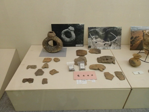 発掘時の3枚の写真が立てかけられたボードや欠片をつなぎ合わせた土器や壺などが展示されている写真