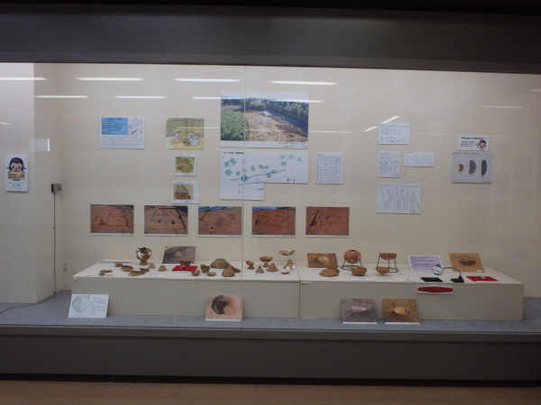 後方の壁に発掘場所の写真などが貼られ、手前に発掘時の写真ボードと壺やお皿などが並んでいる写真
