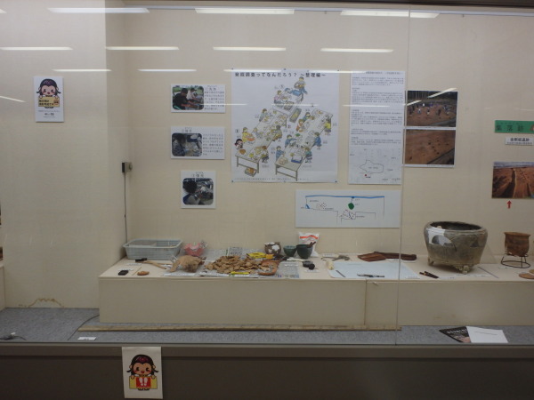 後方の壁に発掘調査の仕事の内容が貼られ、展示台に出土品の欠片などが展示されている写真