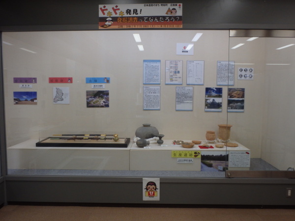 後方の壁に写真や資料が貼られ、手前の展示台に様々な形の壺や2本の刀などが展示されている写真