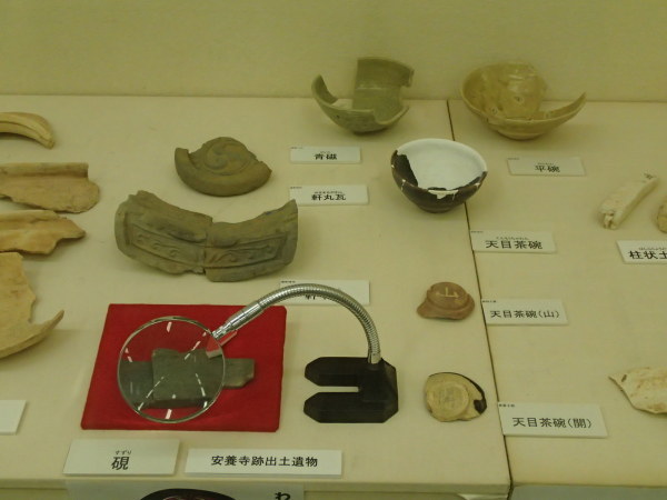 「安養寺跡出土遺物」と書かれた展示台に天目茶碗や欠片、青磁や手前に拡大鏡が設置された硯が置かれている写真