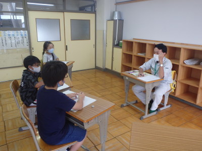 職員の男性と班に分かれた生徒の皆さんが、向かい合うように机を置いて席に着き、生徒から取材を受けている様子の写真