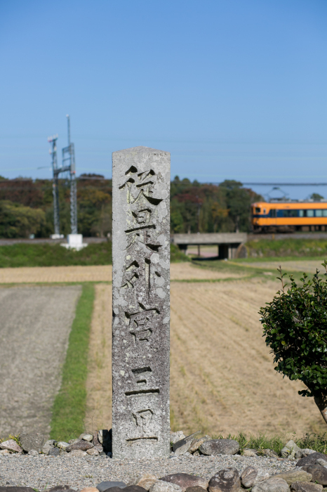 三里の道標とその奥に黄色い電車が走っている風景写真