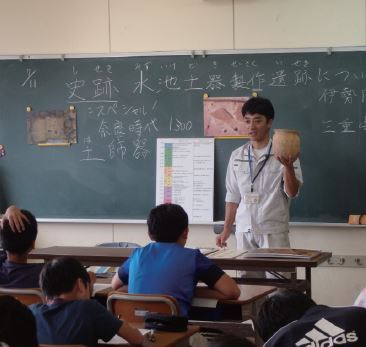 教室の黒板の前で、職員の男性が土器を左手に持って、子どもたちに授業を行っている様子の写真