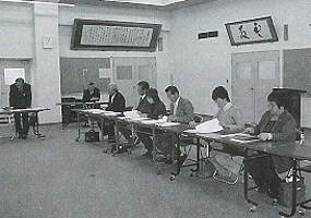 右側に横一列に設置された長机の席に議員の方々が座り奥の席の男性が席を立って意見を述べている会議の様子の白黒写真