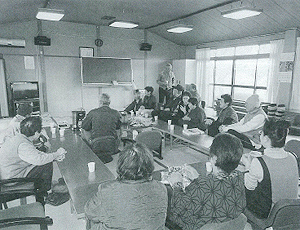 コの字に設置された長机の席に座っている参加者が前方を見ている白黒写真