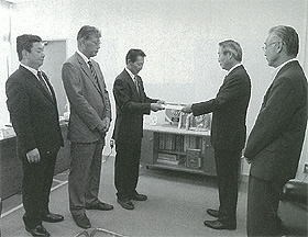 左側に3名右側に2名の男性が向かい合って立ち奥の2名の男性が報告書を手渡ししている白黒写真