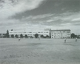 3階建ての校舎が建つ手前の校庭で体育の授業を行っている明和中学校の白黒写真