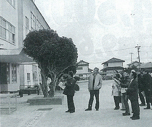 左側の3階建ての校舎の前で議員の方々が校舎を見上げている白黒写真