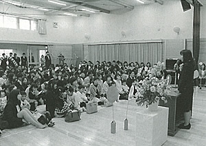右側の檀上に立っている女性の話を親子が座って聞いている入園式の様子の白黒写真