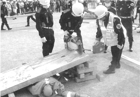 ヘルメットを被った3名の消防団員が機械を使って建物の下敷きになったマネキンを助けている防災訓練の様子の白黒写真