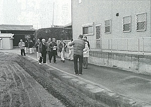 右側の建物と左の畑の間の細い通路で参加者が手前の男性が話を聞いている現地調査の様子の白黒写真