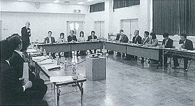 コの字に設置された長机に参加者が席に座り左奥に立っている男性が発言を行っている白黒写真