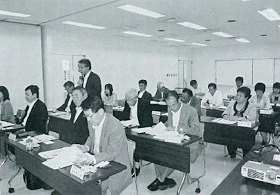 2名ずつ長机の席に座っている参加者の前から2列目の男性が立ち意見を述べているのを聞いている白黒写真