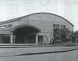 モニュメントが設置されている奥に大きな入り口で屋根が弯曲に設計された総合体育館の建物外観の白黒写真