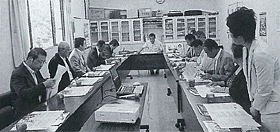 コの字に設置された長机の席に参加者が座り右手前に立っている女性が立ち意見を述べている白黒写真
