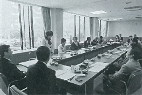 ロの字に設置された長机の席に参加者が座り左奥に立っている女性の話を聞いている白黒写真