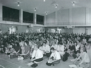 防災講習会の参加者が体育館の床に座り前方を見ている白黒写真