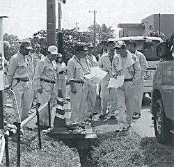三角コーンが置かれている道路の左側の側溝部分蓋の上に作業着を着た議員の方々が集まり調査を行っている様子の白黒写真