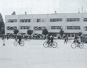 3階建ての校舎の前のポールが置かれた校庭で生徒たちが一人ずつ自転車に乗り安全運転の講習を受けている白黒写真