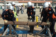 校庭で2名のヘルメットを着用した消防団員が丸太の端を支え中央の男性が垂直にカットしている写真