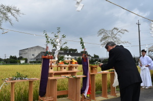 黄金色に稲穂が実った田んぼの前に、祭壇が設けられ、スーツ姿の男性が祭壇前で、頭を下げており、神事が行われている様子の写真