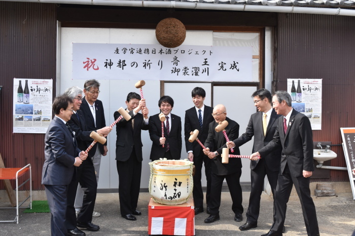 「祝 神都の祈り  斎王 御裳濯川 完成」と書かれた横断幕が掲げられ、完成イベントで、関係者の方々8名が木づちを持ち、樽酒で鏡開きが行われている様子の写真